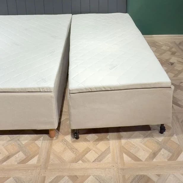 Zwei fertig montierte Bettmodule stehen mit einem schmalen Spalt nebeneinander auf einem hellen Holzboden. Jedes Modul hat einen eigenen beigen Bezug, eine eigene gesteppte Matratze und eigene Beine.
